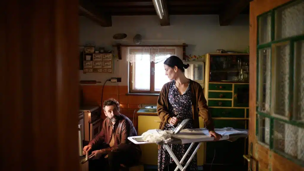 A woman is ironing in a rustic kitchen while a man sitting nearby looks towards her.