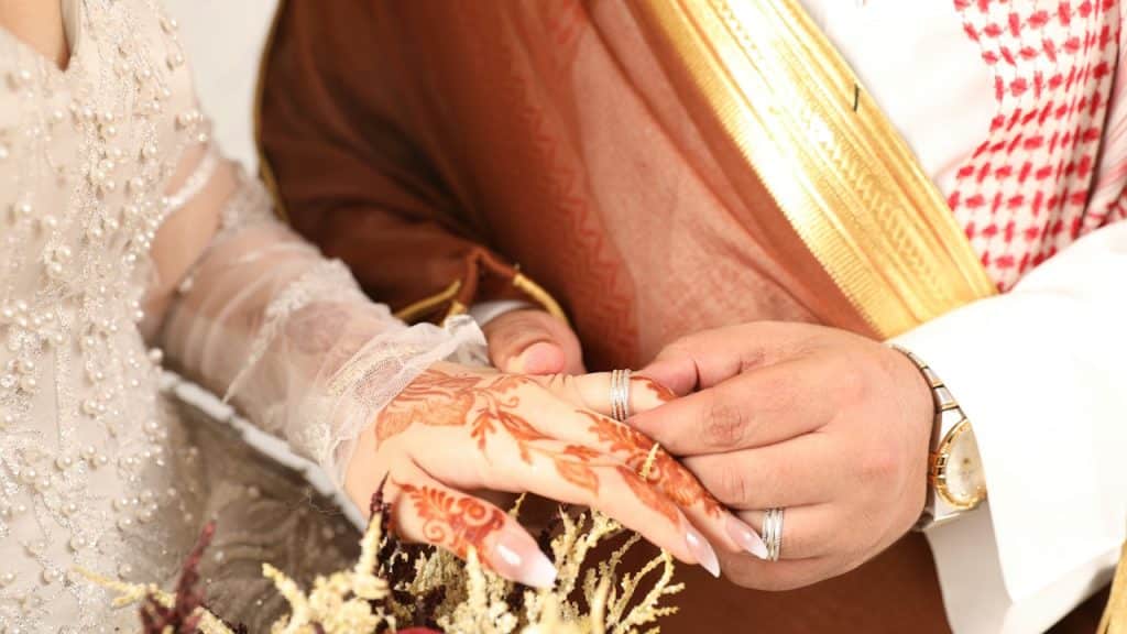Close-up of a groom placing a ring on a bride's hand, which is decorated with henna, while both wear traditional Arab wedding attire.