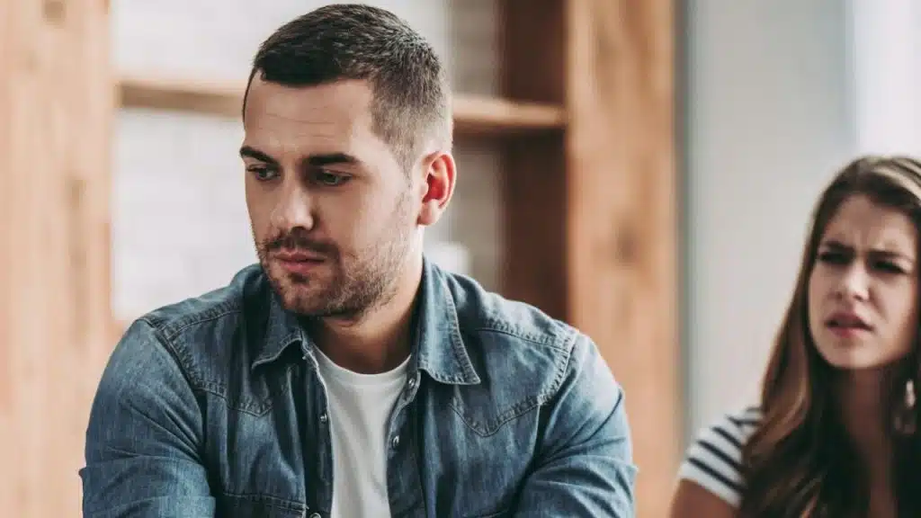 A man with a serious expression is looking down while a frustrated woman is blurry in the background.