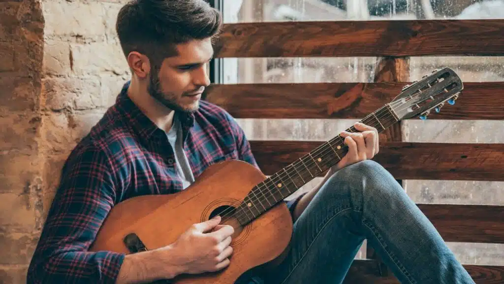 Man in a plaid shirt sitting by a window and playing an acoustic guitar.