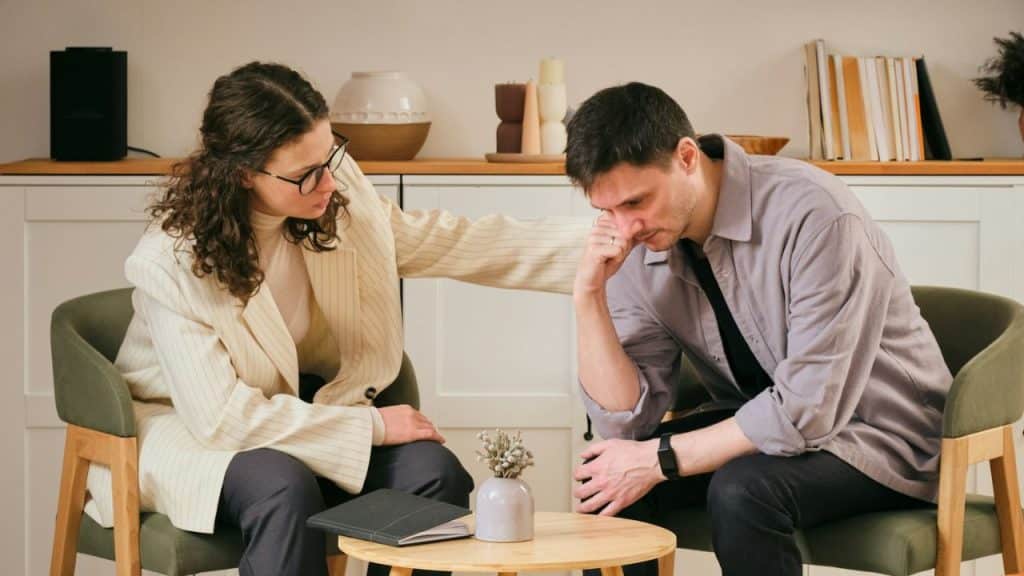 A woman in glasses comforts a man with his head in his hand in a modern office.