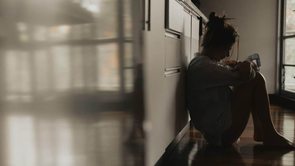 A woman is sitting on the floor, leaning against a dark cabinet, hugging her knees.