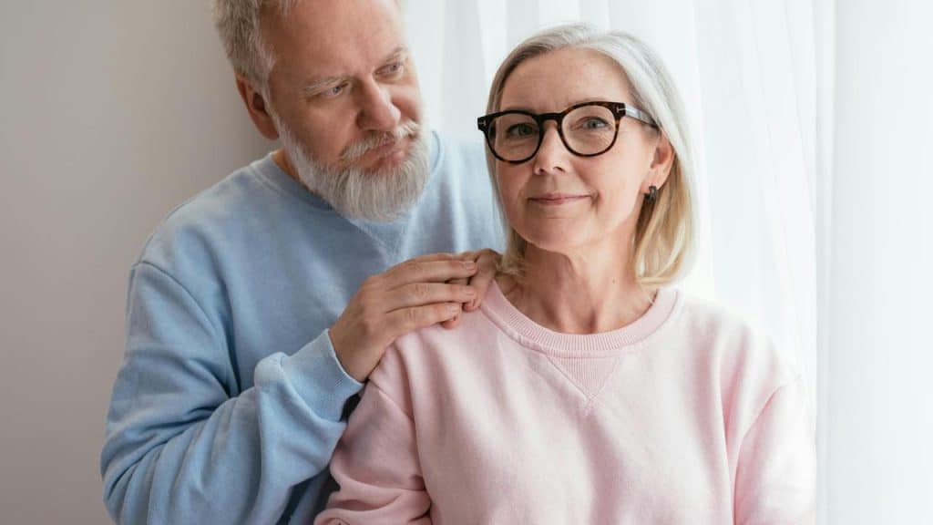 An elderly man gently placing his hands on a womanโs shoulders as she smiles softly.