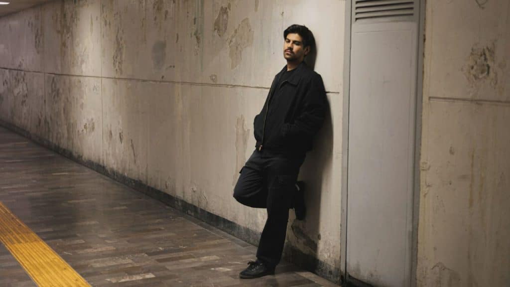 A man in dark clothing leans against a weathered concrete wall in a hallway.