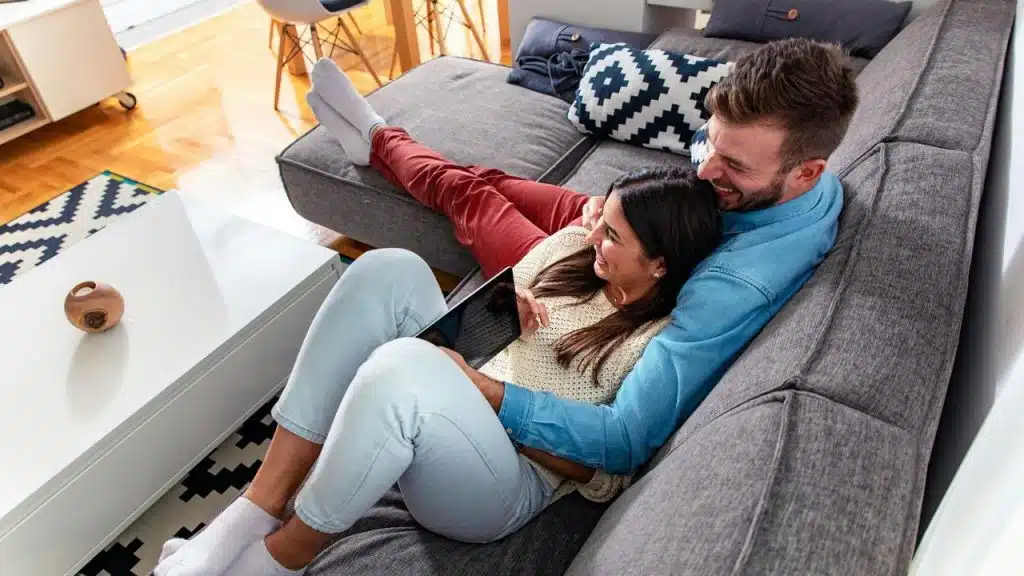 A smiling couple cuddling on a sofa while looking at a tablet together.
