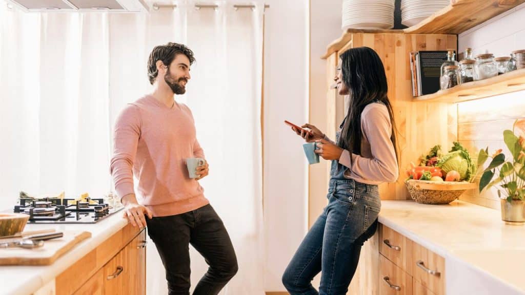 A man and a woman standing in a kitchen, talking and holding mugs.