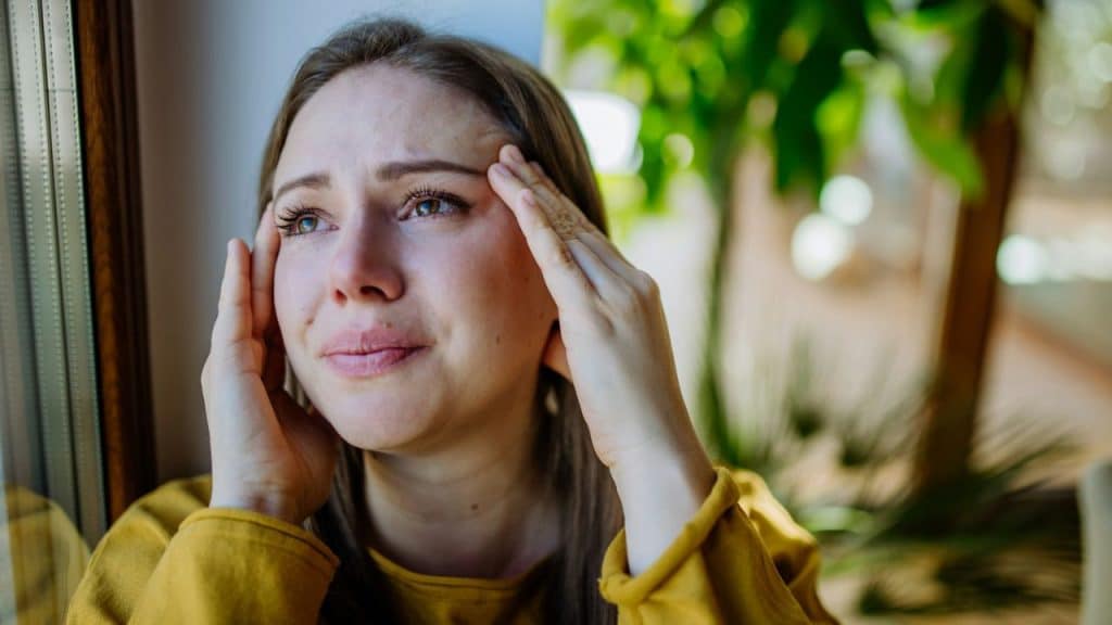 Crying woman in a yellow shirt with hands on her temples looking out a window in a sunlit room.
