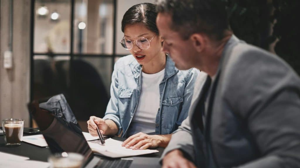 Woman wearing glasses and denim shirt pointing at a document to a man in a blazer.