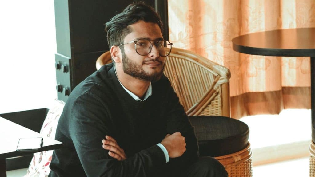 Man with a beard and glasses sitting with his arms crossed in a wicker chair.