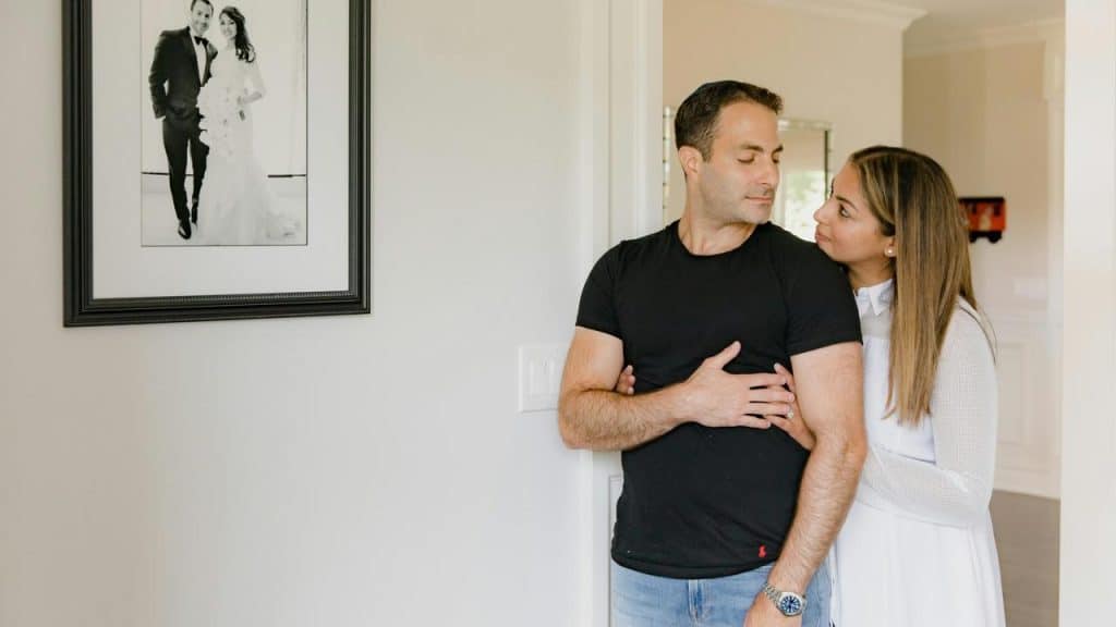 A woman embraces a man from behind as they stand near a framed wedding photo on the wall.