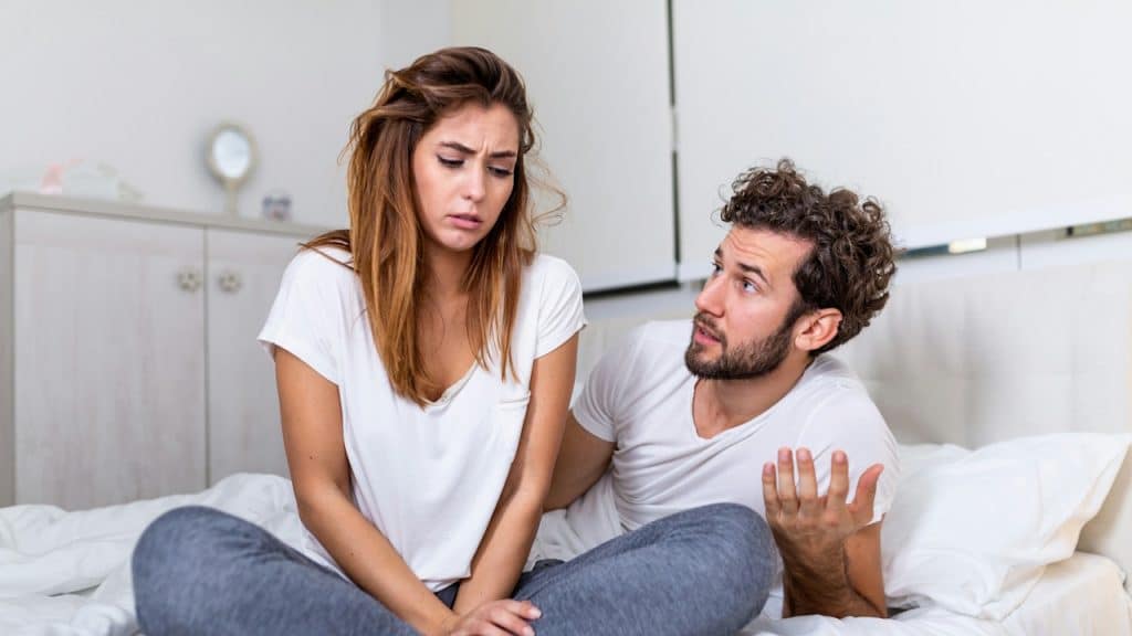 A distressed woman is sitting cross-legged on a bed while a man next to her is gesturing and speaking intently to her.