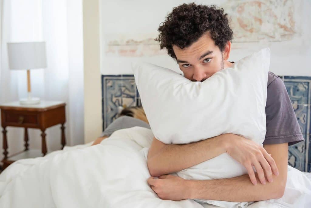 The sad man is hugging the pillow while the woman is sleeping beside him.