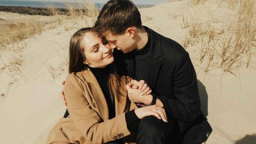 A couple sitting close together on sand dunes, holding hands.