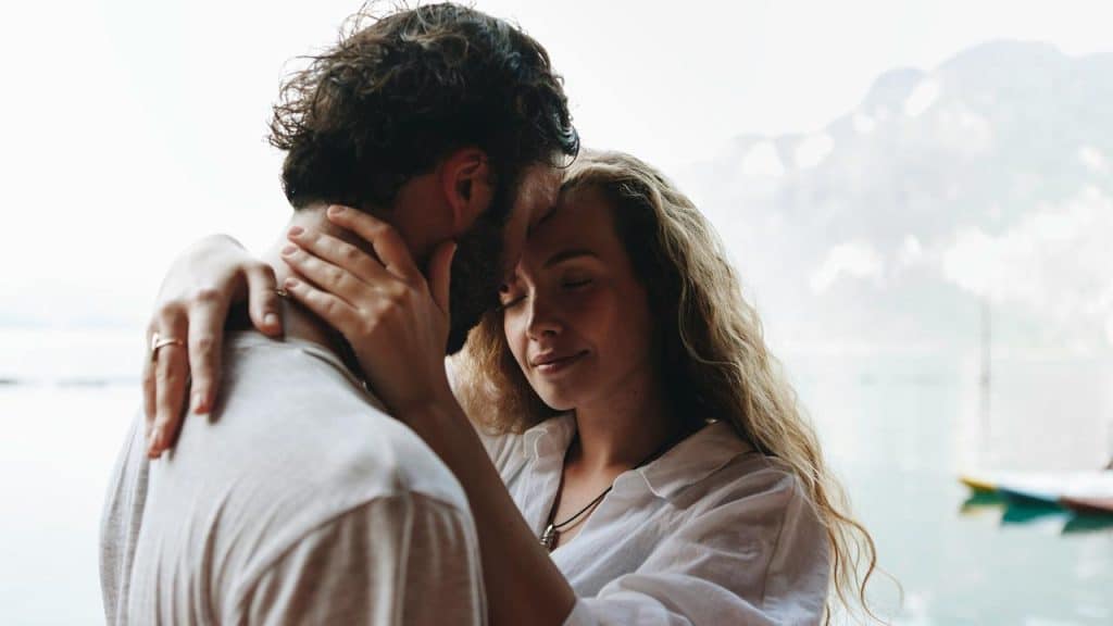 A couple shares an intimate moment by a calm lake with mountains in the background.