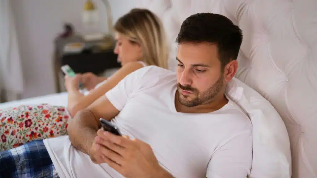 A man and a woman lie in bed, separately focused on their phones, facing away from each other.