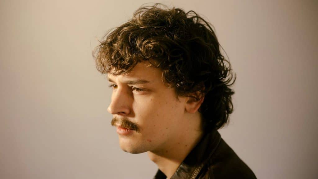 Profile view of a young man with curly hair and a mustache against a beige background.