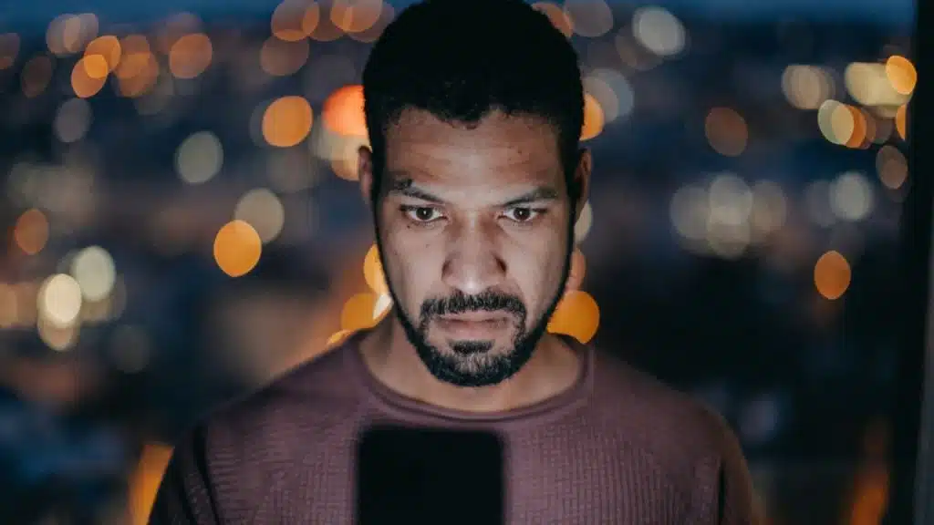 A serious man with a beard looks down at a phone with blurry city lights in the background.