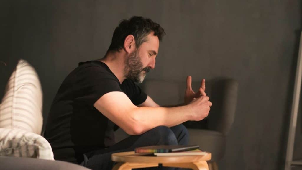 A bearded man sits with his hands clasped, looking down thoughtfully next to a small wooden table.