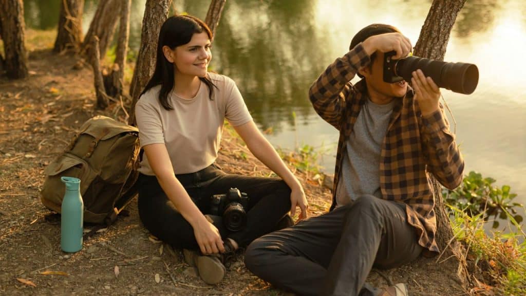A woman smiles as she sits with a camera next to a man taking a photo with a telephoto lens near a body of water.