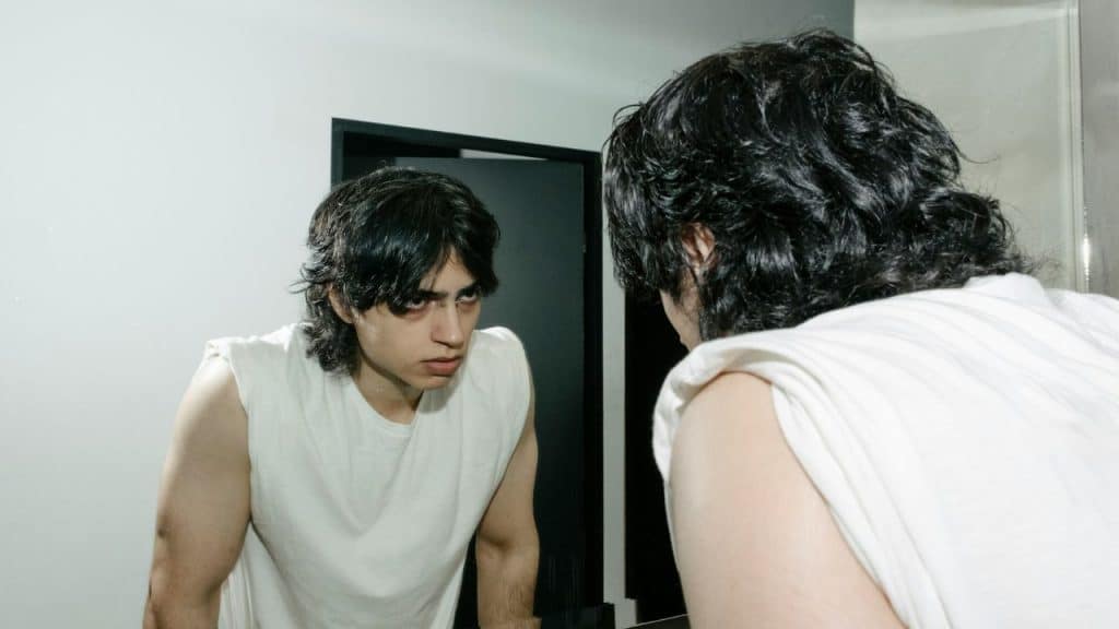 A young man with dark, curly hair and a white sleeveless shirt stares intently at his reflection in a mirror.