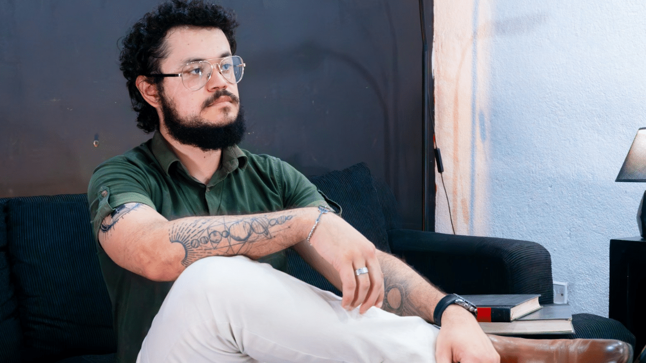 Man with glasses and a beard sitting on a couch, showing arm tattoos.