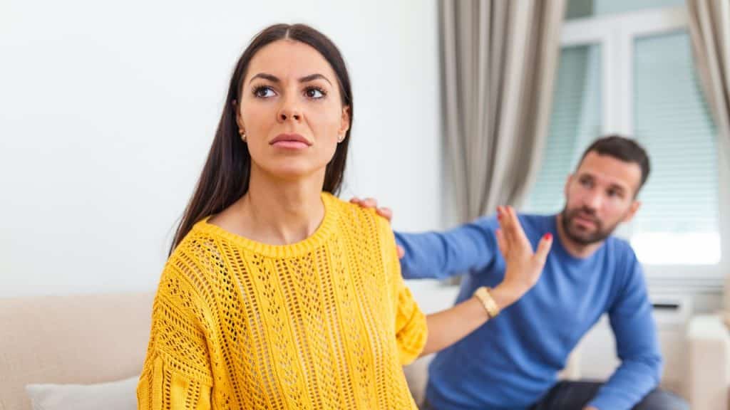 A woman in a yellow sweater is turning away from a man in a blue shirt who is trying to touch her shoulder.