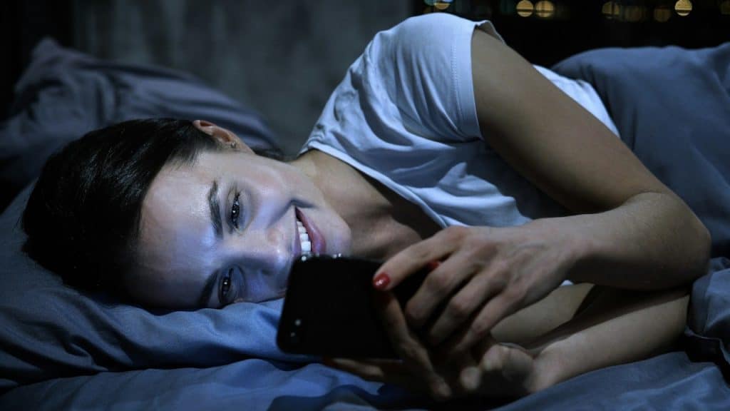 A smiling woman lies in bed in the dark, looking at her smartphone.