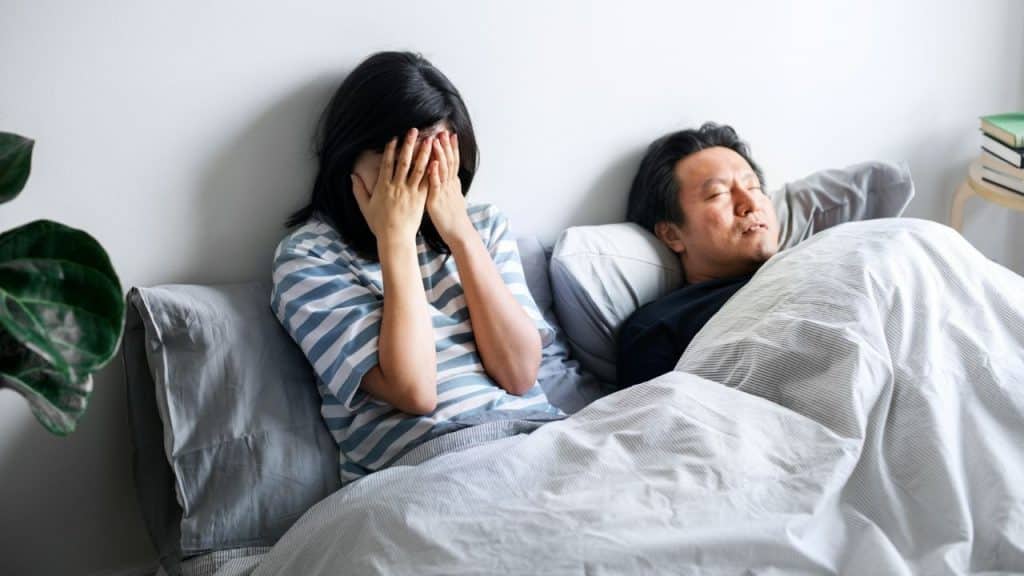 Woman crying into her hands while a man sleeps next to her in bed.