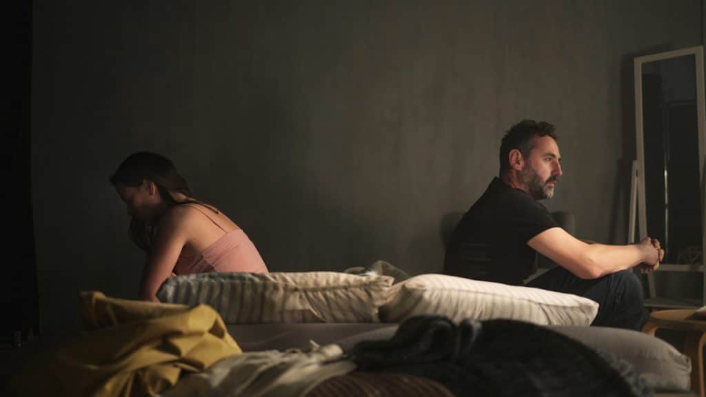 A man and a woman sit facing away from each other on a bed in a dimly lit room, appearing distant and troubled.
