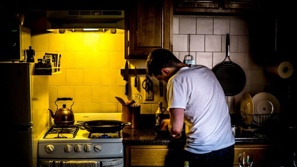 A man in a white T-shirt is cooking in a dark kitchen, illuminated by the bright, yellow light of the stove area.