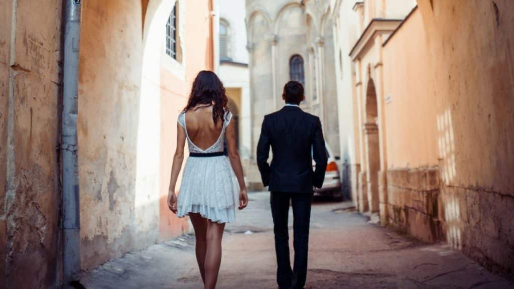 A couple in formal clothes walks away down a narrow, old European street.