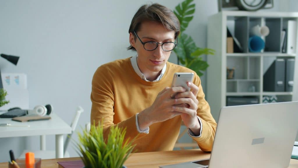 A focused young man in a yellow sweater and glasses, sitting at a desk, looking down at and using his smartphone with a laptop nearby.
