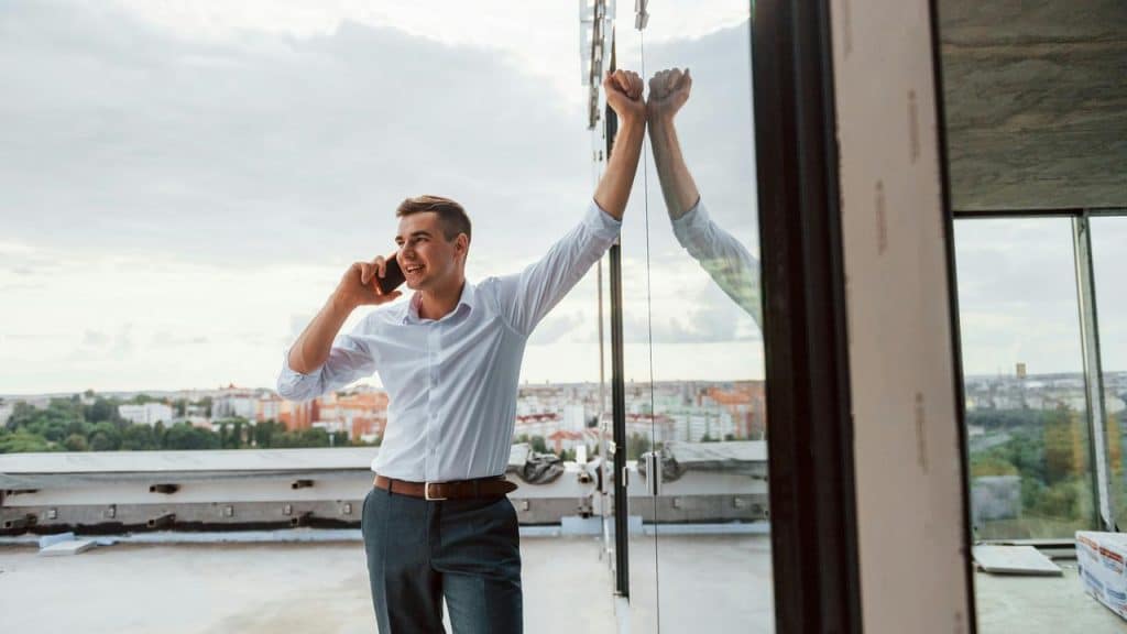 A man in a dress shirt talking on the phone by a glass wall with a city view.