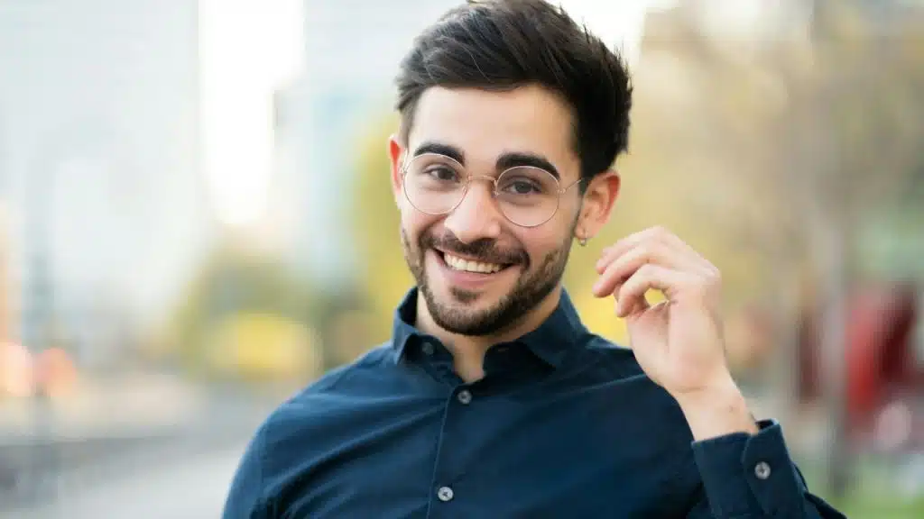 Smiling young man with a beard and round glasses posing outdoors.