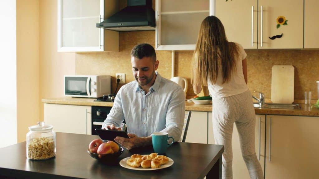 A man is sitting at a kitchen table looking at a tablet while a woman with long hair stands at the counter, preparing food.