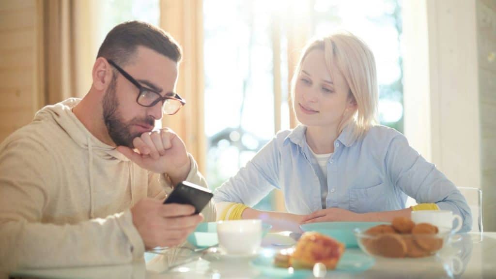 A man intently checks his phone at a table while a woman watches him with a subdued expression.