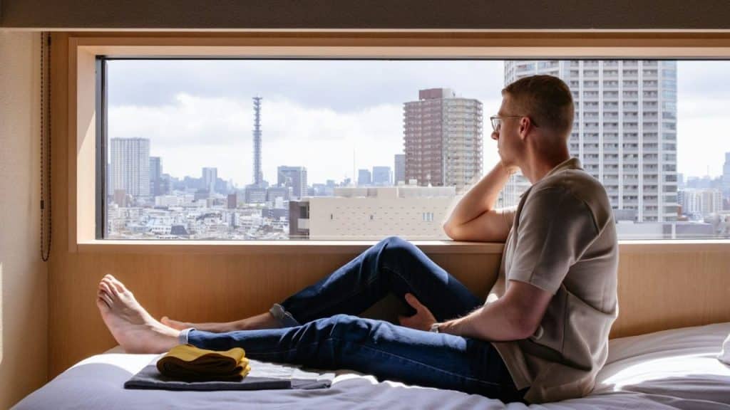 A man sits on a bed, looking out a large window at a city skyline.