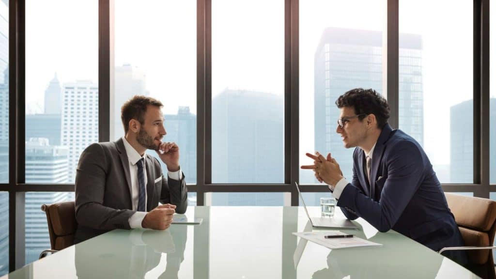 Two men in suits talking across a table in a high-rise office with a city view.