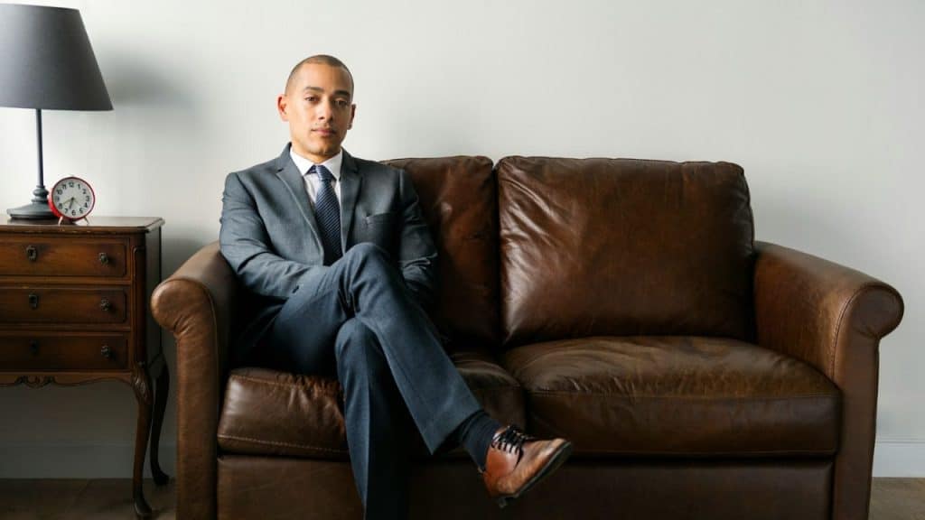 Man in a suit sitting formally on a brown leather couch looking directly at the camera.