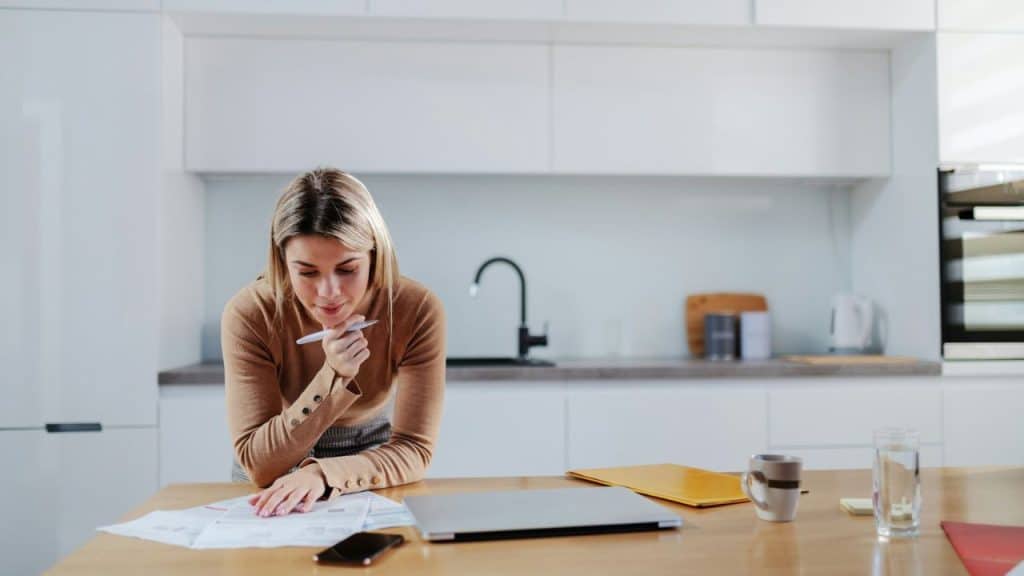 Woman with pen reviewing papers on a kitchen counter with a laptop and phone.