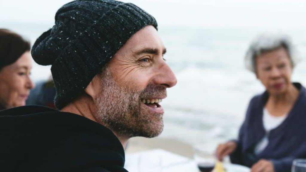 Middle-aged bearded man in a knit cap smiling outdoors near other people.