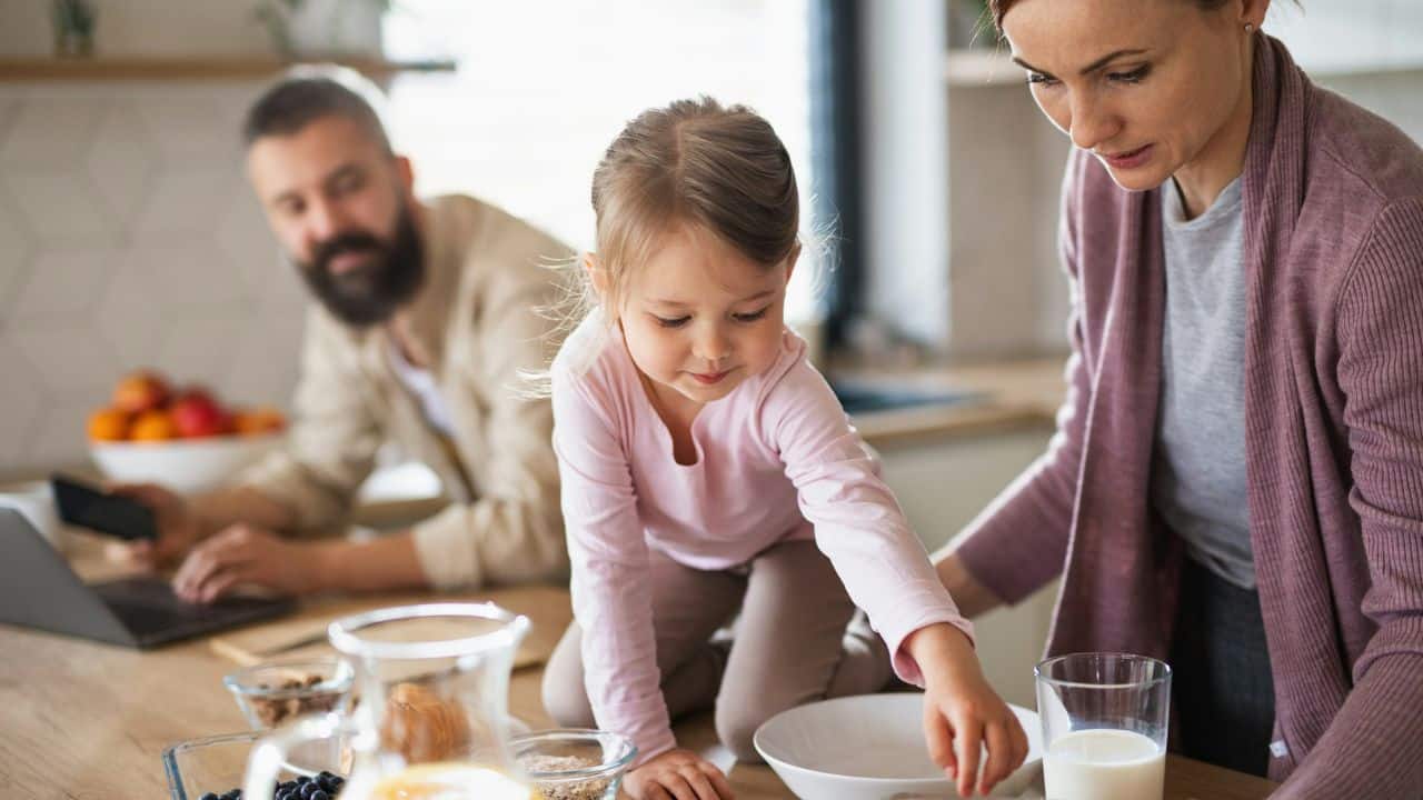 Mother and young daughter preparing food at a kitchen counter with a father blurred in the background.