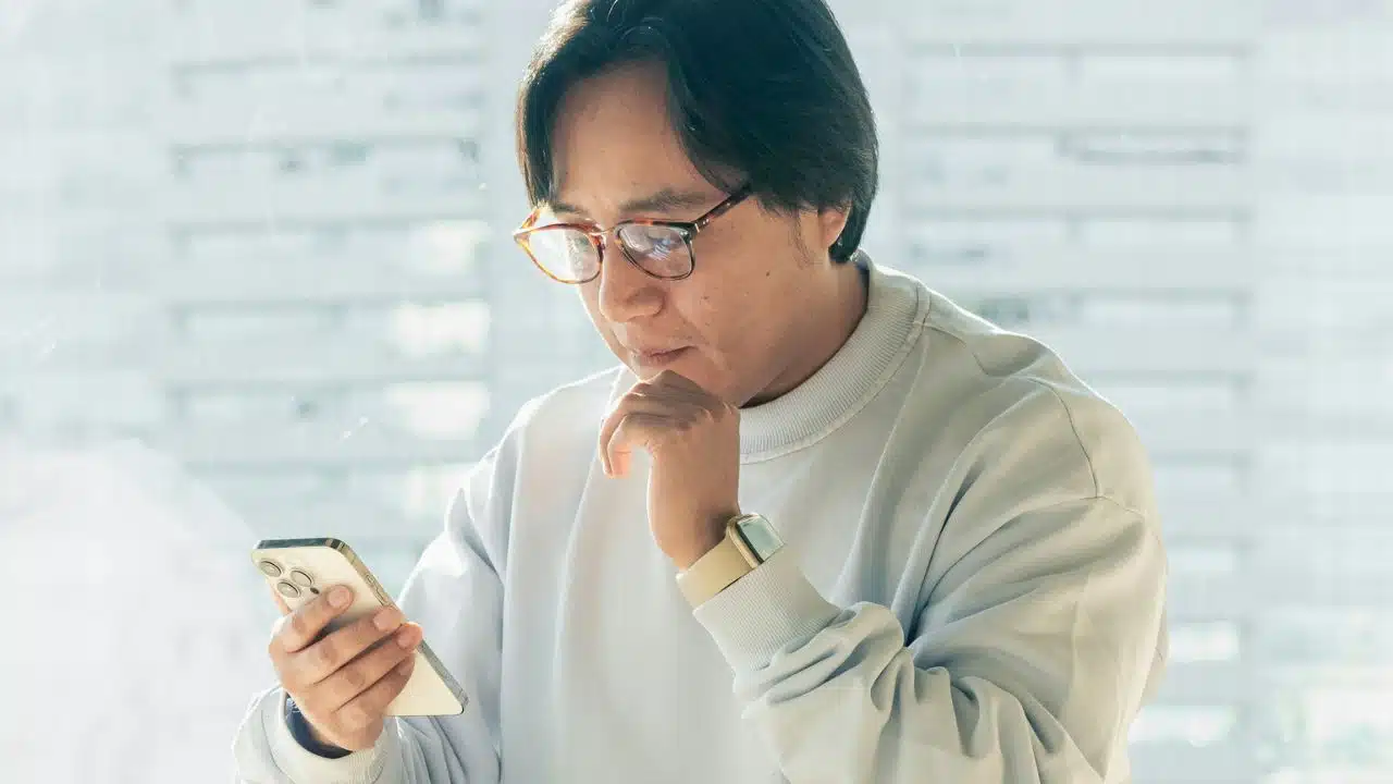 Man with glasses and a light sweatshirt looking intently at a smartphone in his hand.