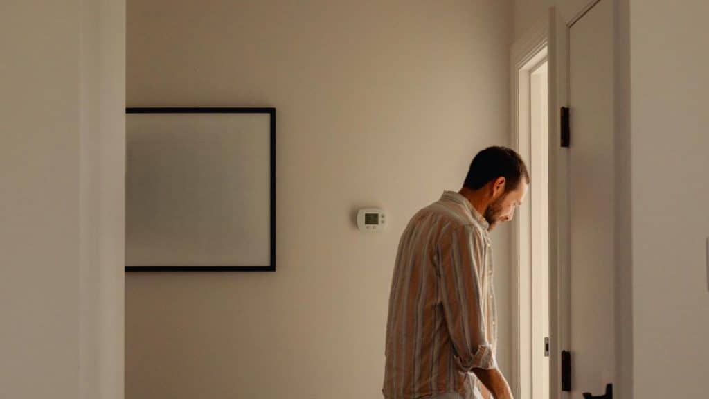 A man standing near a doorway in a softly lit room, looking down thoughtfully.
