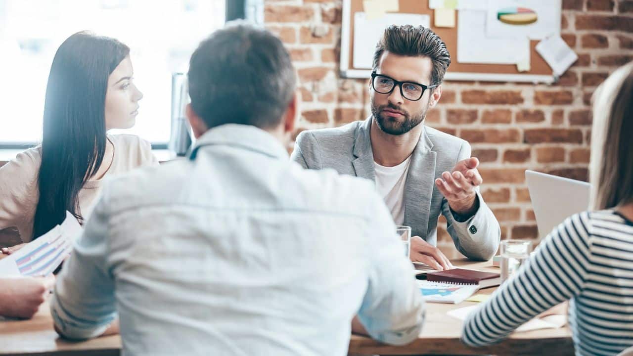 Man in glasses and a gray blazer speaks to a team around a conference table.