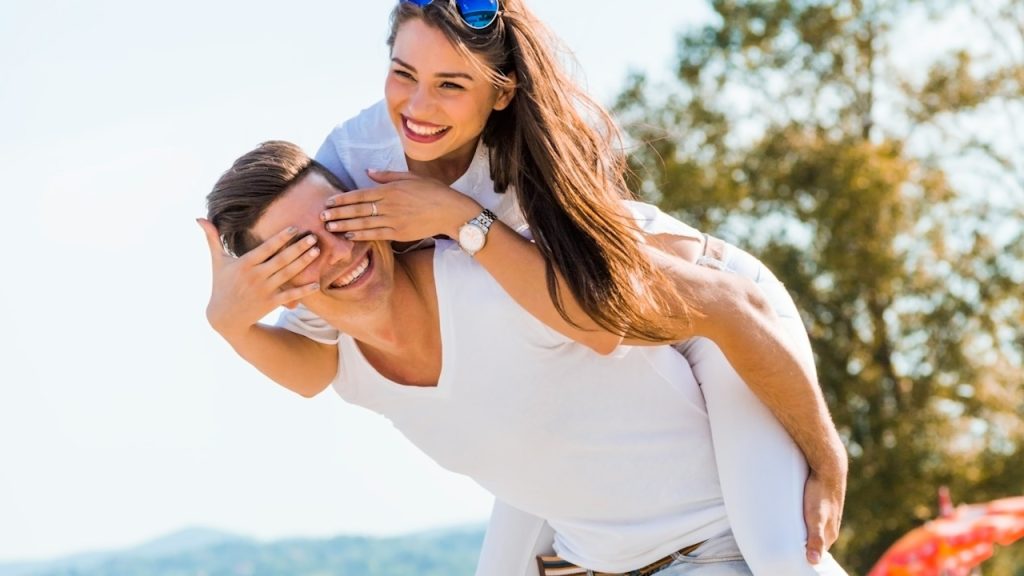 A man carrying a woman on his back while she covers his eyes with her hands.