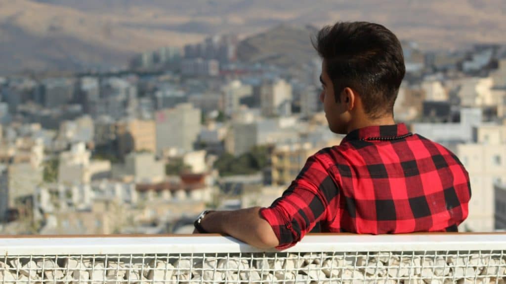 A man wearing a red checkered shirt sitting on a bench overlooking an urban area.