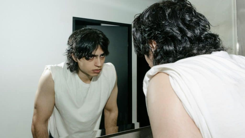 A young man with dark, long hair in a white tank top intensely stares at his reflection in a mirror.
