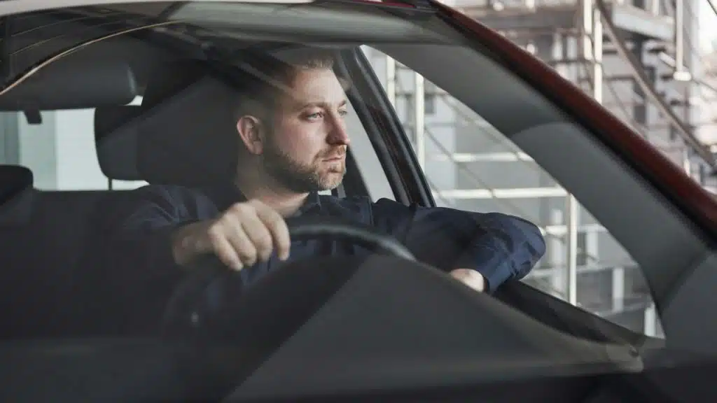 Bearded man in a dark shirt driving a car, looking out the side window.