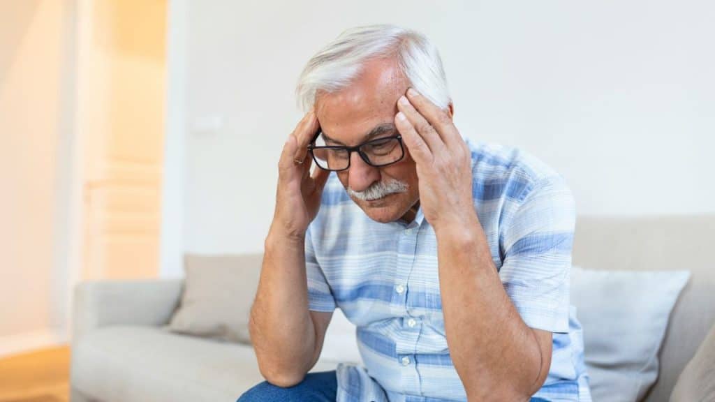 Elderly man with glasses and mustache, sitting and holding his temples in distress.